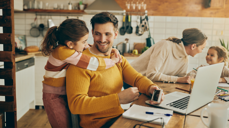 Person in a mustard sweater sits at a wooden table with a laptop and papers, while a child hugs them from behind; another adult and child chat in a kitchen.