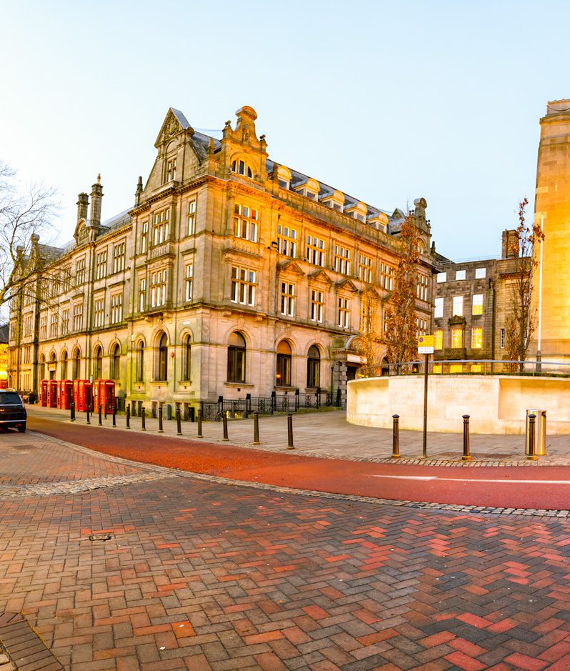 A panoramic view of a city square at dusk featuring historic buildings, leafless trees, street lamps, and cobblestone streets with some cars parked nearby.
