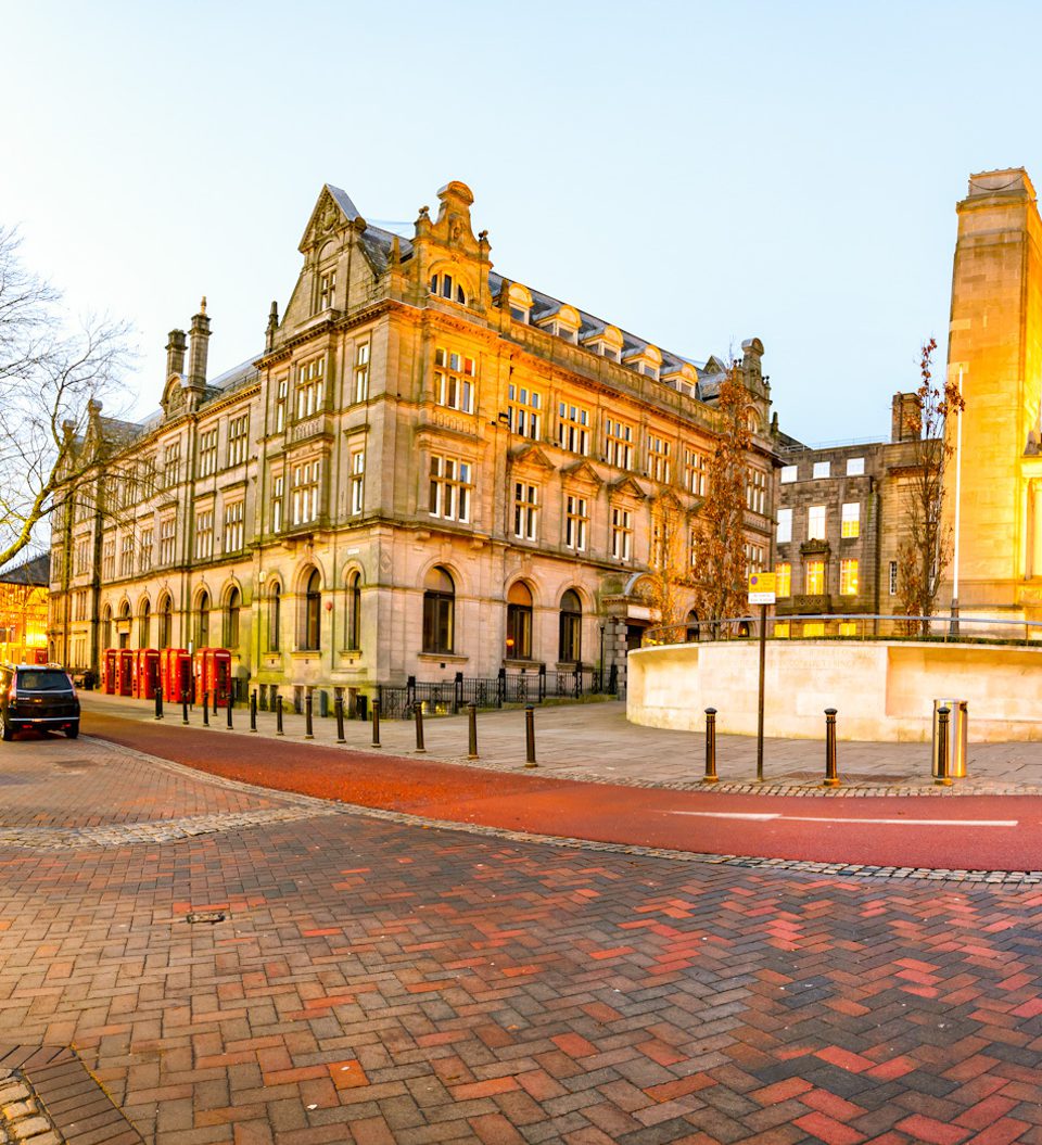 A panoramic view of a city square at dusk featuring historic buildings, leafless trees, street lamps, and cobblestone streets with some cars parked nearby.