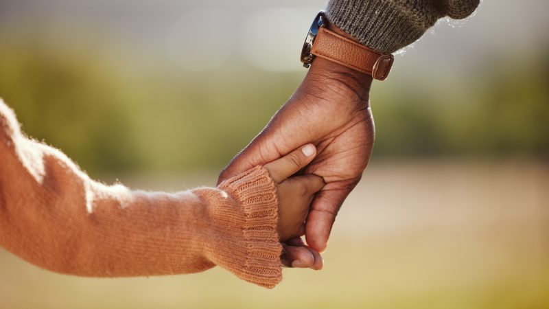 A close-up of an adult and child holding hands outdoors, with the child's hand gently grasping the adult's finger. The adult wears a brown leather watch and a grey knitted sweater, while the child wears a cosy orange sweater. The background is blurred with greenery.