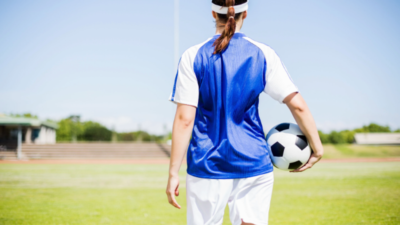 Back view of a person in a blue and white football jersey, holding a black-and-white football on a sunny grassy field with stands in the distance.