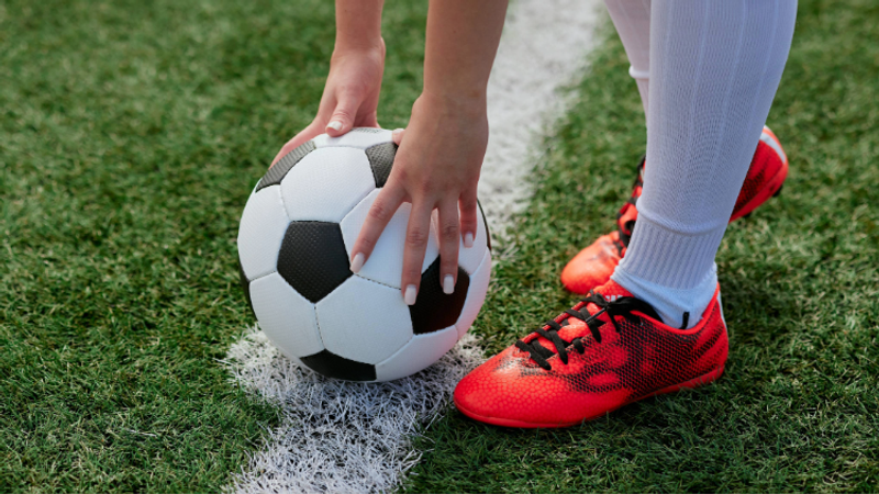 A person in football boots is about to kick a black-and-white soccer ball on a grass field, with hands gripping the ball and a white line nearby.