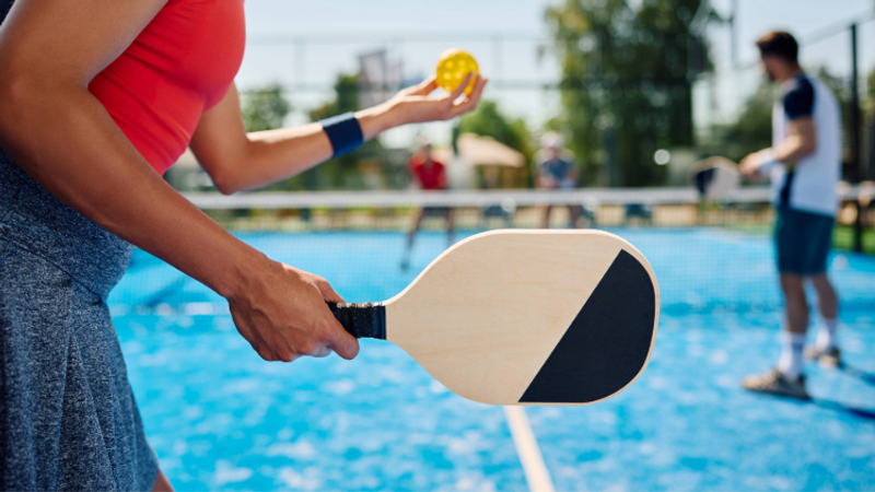 A person holding a paddles, with a yellow ball in their hand, on a blue pickleball court, with other players in the background.