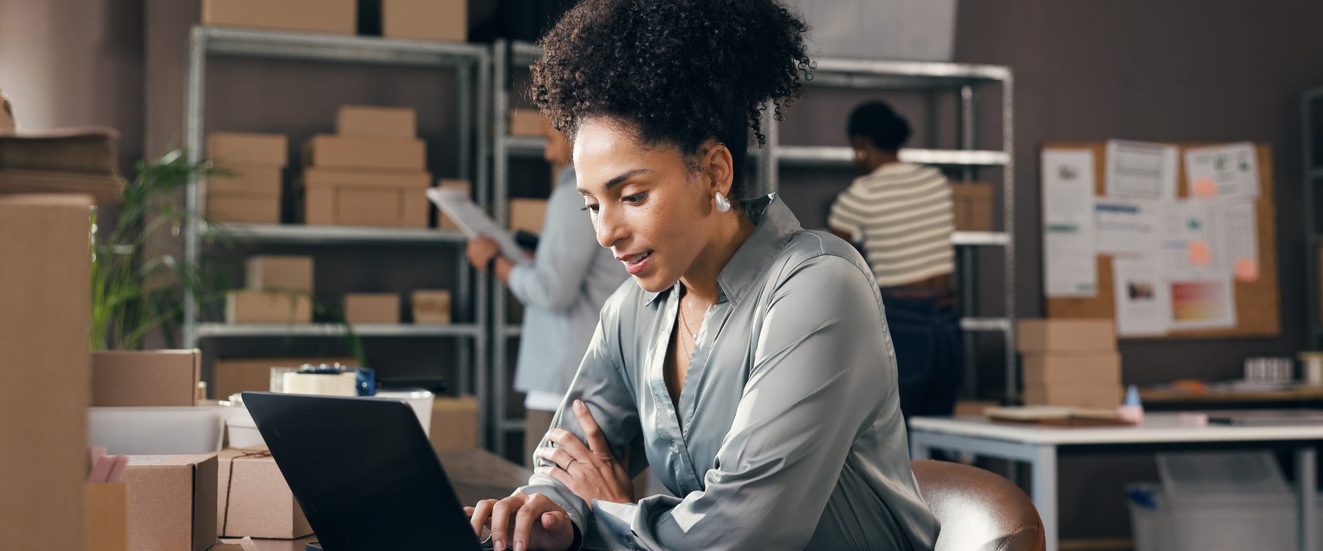 Person with curly hair sits at a desk with a laptop in a busy storage workspace, surrounded by boxes, shelving and other staff in the background.