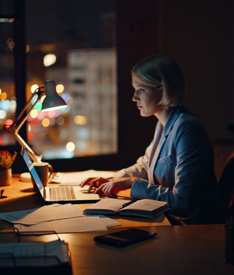 A woman in a blue blazer working on a laptop at a cluttered desk in a dimly lit office, with city lights visible through the windows behind her at night.