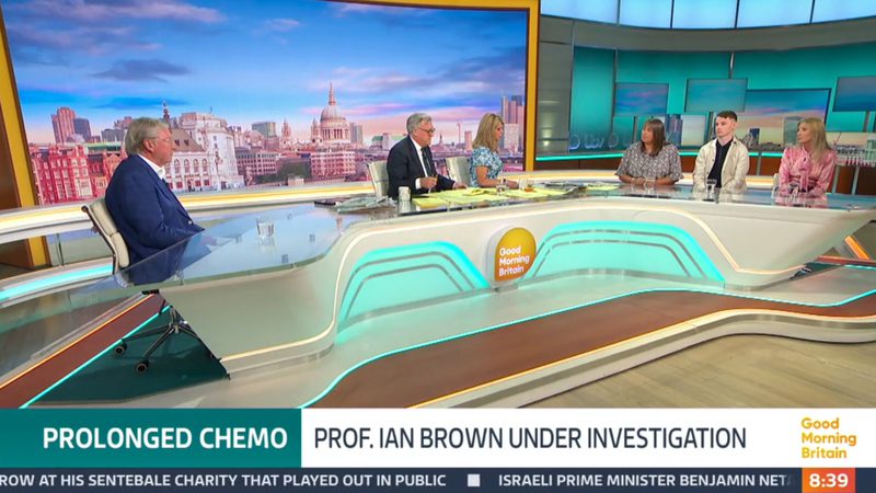A television studio with six panel members seated at a curved desk, a cityscape background on screens, and a "Good Morning Britain" logo on the desk. The time displayed is 8:39.