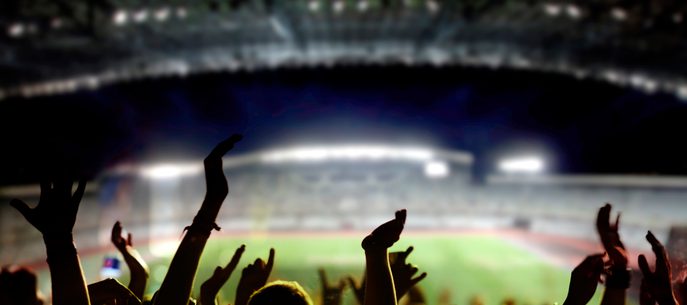 Silhouetted crowd with raised hands in a packed stadium at night, bright stage lights creating a hazy glow as the arena and pitch blur in the background.