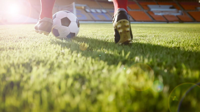 Low-angle close-up of a football on green grass as a player's cleats near the ball, bright sun to the left and empty stadium stands in the background.