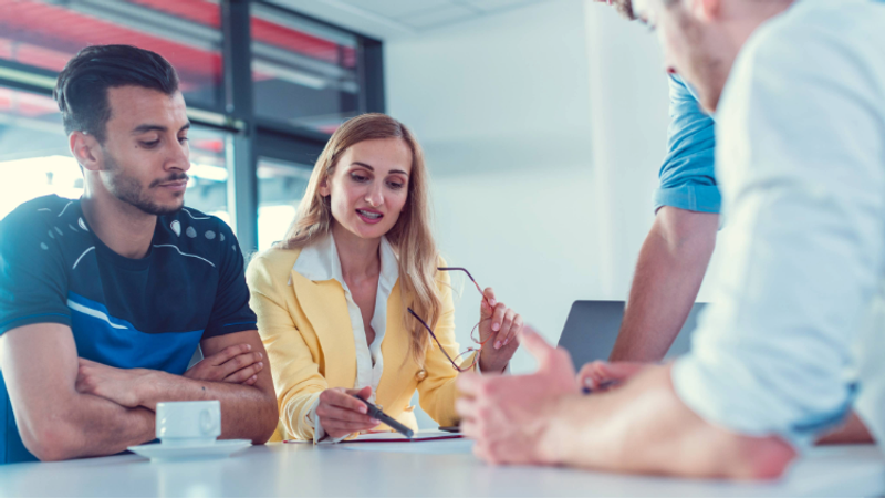 A woman in a yellow blazer discusses with a man while holding glasses, with two other people partially visible in a bright, modern office.