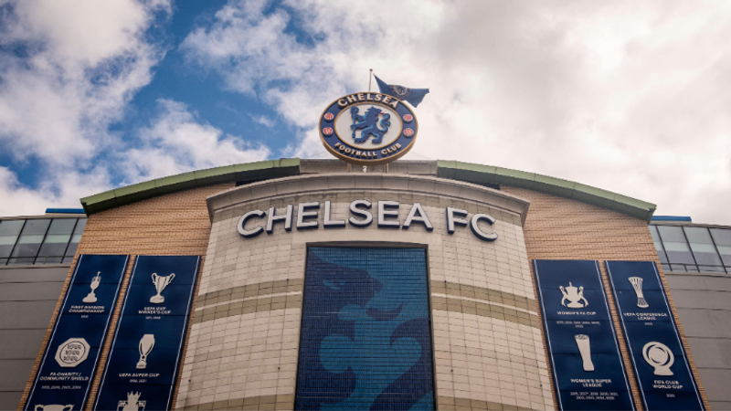 Chelsea FC stadium facade with a curved sign reading CHELSEA FC, a crest atop, and blue banners displaying trophies against a cloudy sky.