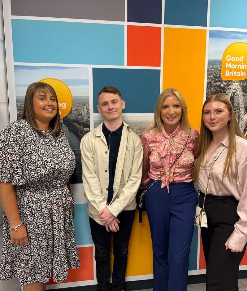 Four people standing in front of a colourful wall with "Good Morning Britain" logo, smiling and dressed in casual and smart clothing.