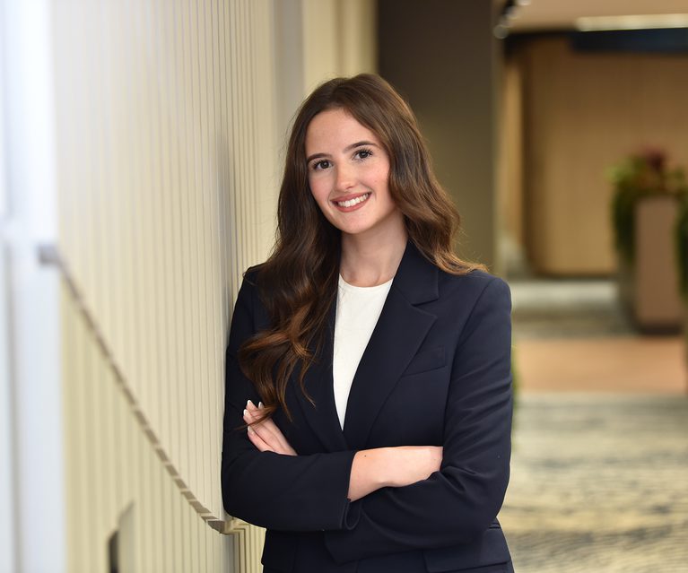 A young woman with long wavy brown hair, wearing a navy blue blazer and white top, smiling confidently while standing with her arms crossed indoors.