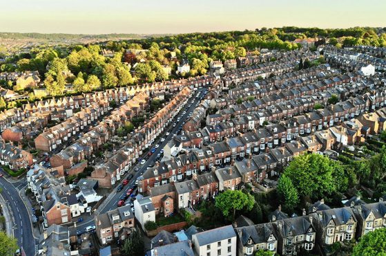 Aerial view of a densely packed residential neighbourhood with rows of terraced houses, surrounded by green trees and rolling hills in the background.