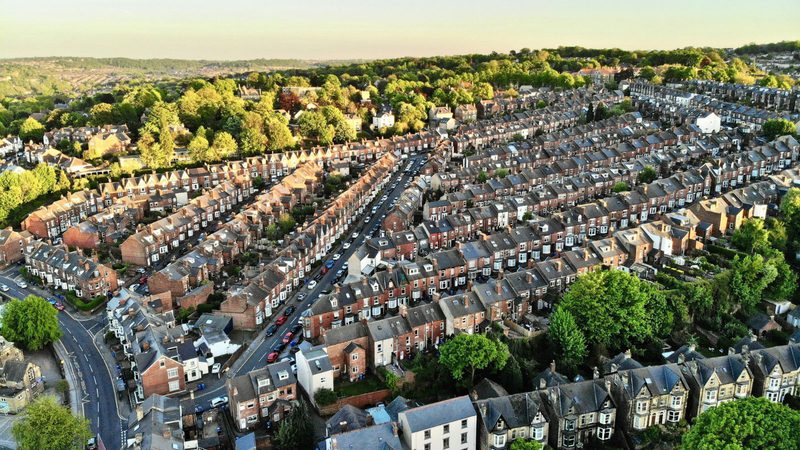 Aerial view of a densely packed residential neighbourhood with rows of terraced houses, surrounded by green trees and rolling hills in the background.