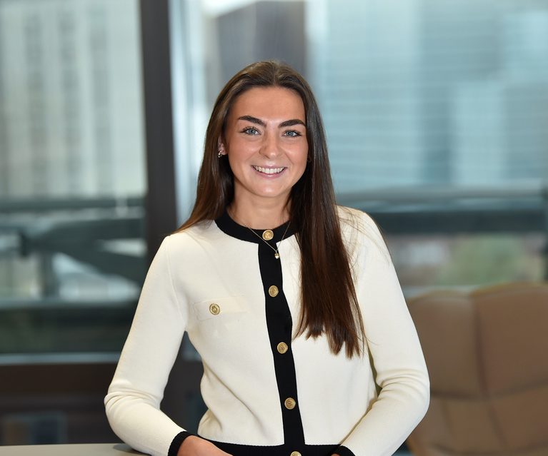 A young woman with long brown hair, wearing a white blazer with black trim and gold buttons, smiling in an indoor office setting with large windows behind her.