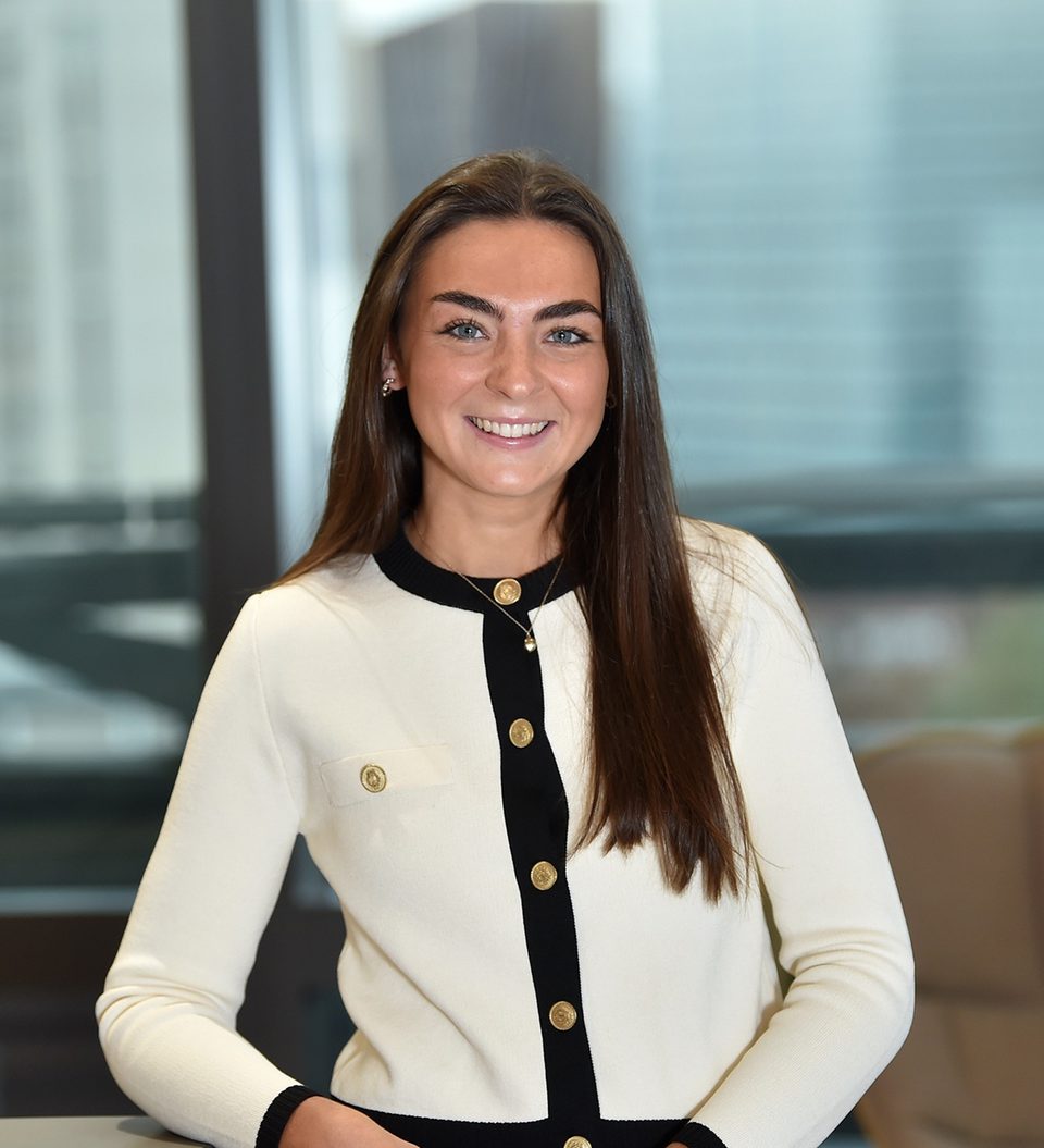 A young woman with long brown hair, wearing a white blazer with black trim and gold buttons, smiling in an indoor office setting with large windows behind her.