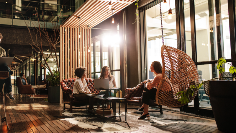 Three women are seated in a modern, well-lit lounge area with large windows, having a conversation around a laptop on a coffee table.