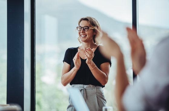 Smiling woman with glasses claps her hands during a meeting in front of a large window, with a blurred person in the foreground.
