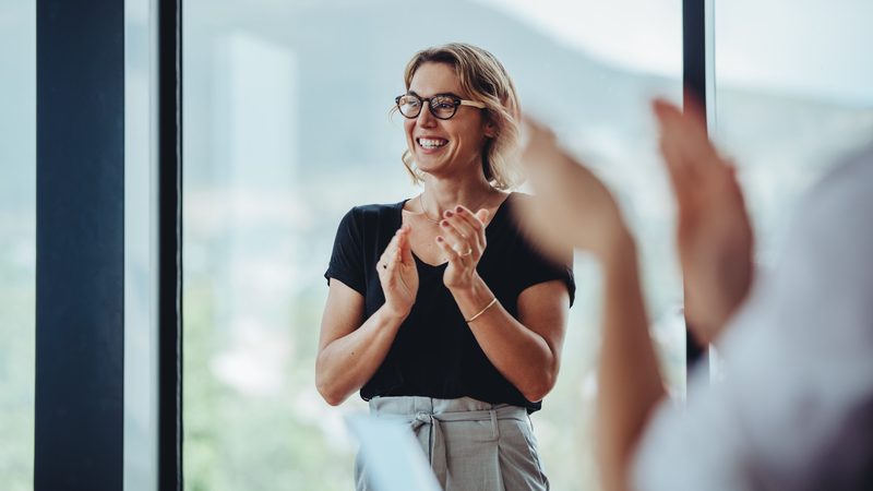 Smiling woman with glasses claps her hands during a meeting in front of a large window, with a blurred person in the foreground.