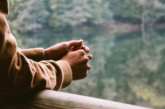 A person wearing a brown sweater holds their hands clasped together over a wooden railing, overlooking a calm, reflective lake surrounded by trees.