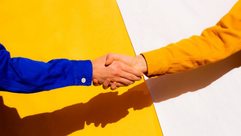 Two people shake hands, one in a blue sleeve and the other in a yellow sleeve, against a bold yellow and white split background with strong shadows.