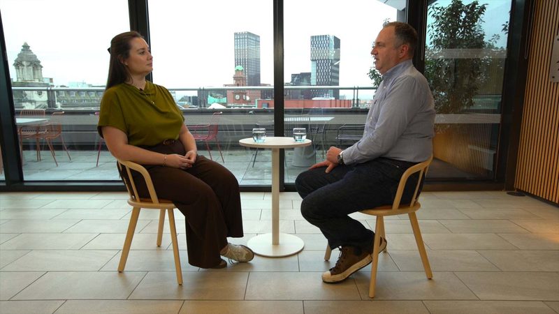 A woman and a man are sitting across from each other in a modern indoor space with large windows, on wooden chairs with a small round table between them. The woman has long brown hair, is wearing a green top and brown trousers, and appears to be listening. The man has short hair, is dressed in a light grey shirt and dark trousers, and is speaking. Two glasses of water are on the table. The background shows a cityscape with various buildings and some outdoor seating on a balcony.