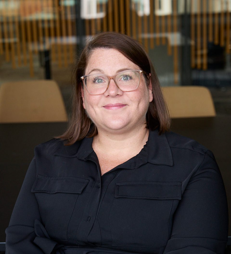 Portrait of a person with shoulder-length brown hair and glasses, wearing a dark shirt, smiling, seated in a modern indoor space with vertical wooden slats.