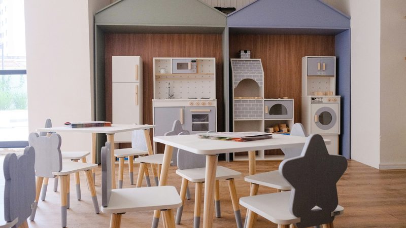 Bright children's playroom featuring a pastel blue and white toy kitchen set, shelves, and appliances; round white tables and small wooden-legged chairs.