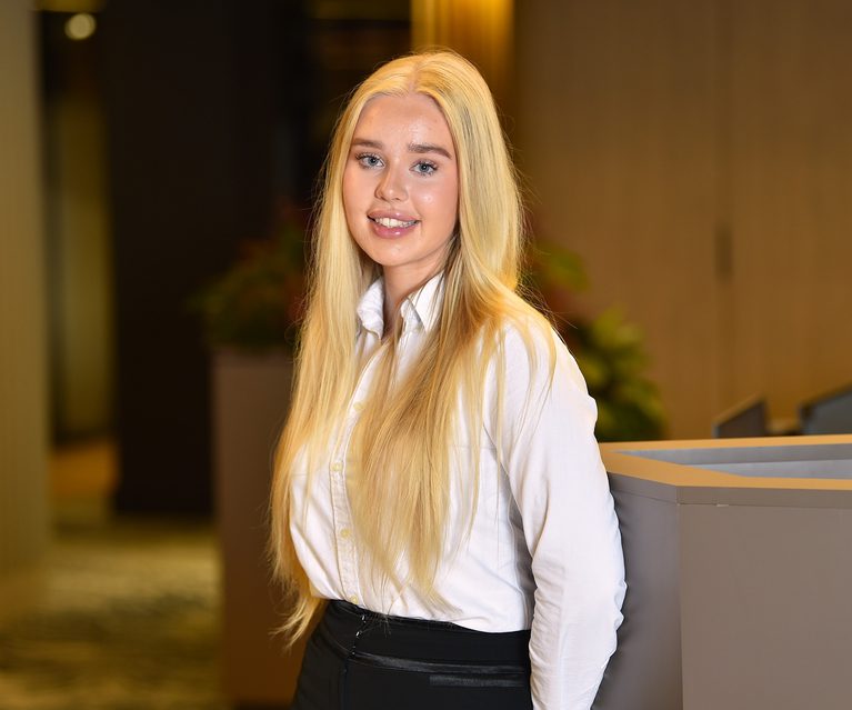 A young woman with long blonde hair, wearing a white shirt and black trousers, standing indoors with a blurred background of warm lighting and plants.