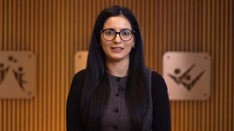 A woman with long dark hair, glasses, and earrings, wearing a grey and black outfit, standing in front of a wooden background with two blurred images behind her.
