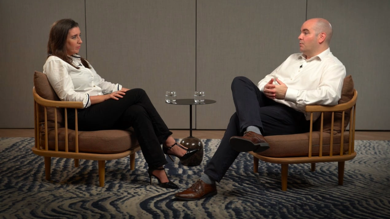 A woman and a man sit facing each other in armchairs with a small round table and two glasses of water between them. The woman has long brown hair and is wearing a white top and black pants. The man has a bald head and is wearing a white shirt and dark trousers.
