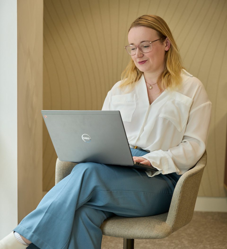 Person with shoulder-length blonde hair and glasses sits in a beige chair, using a Dell laptop on their lap, wearing a white blouse and blue trousers.