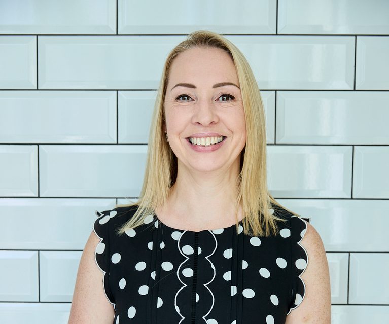 Smiling person with shoulder-length blonde hair wearing a black sleeveless dress with white polka dots and scalloped white edging, standing against pale blue tiled wall.