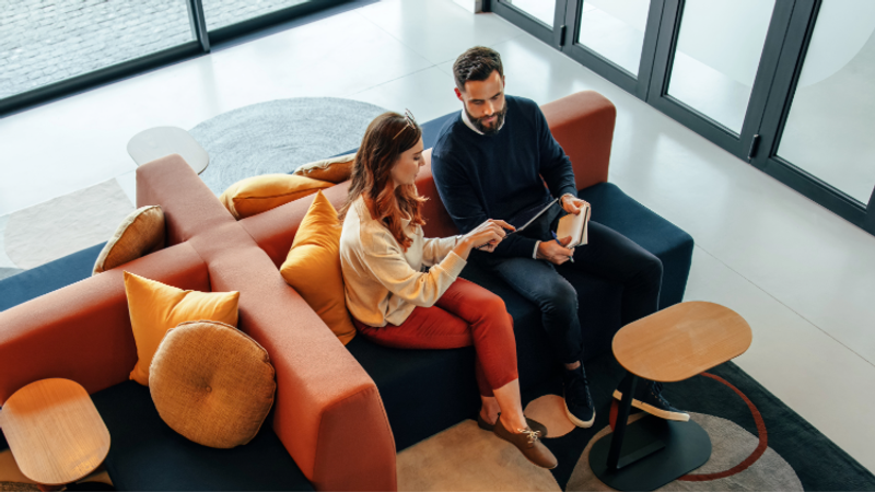A man and woman sit together on a sofa in a modern, brightly lit lounge area, looking at a tablet, with an orange and tan pillow and a small wooden table nearby.