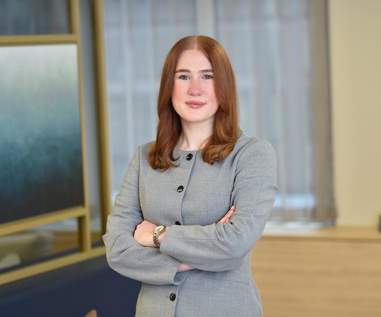 A confident woman with shoulder-length red hair, dressed in a light grey blazer with black buttons, standing with arms crossed in an office setting.