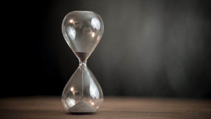 A glass hourglass with black sand flowing from the top chamber to the bottom, set on a wooden surface against a dark background.