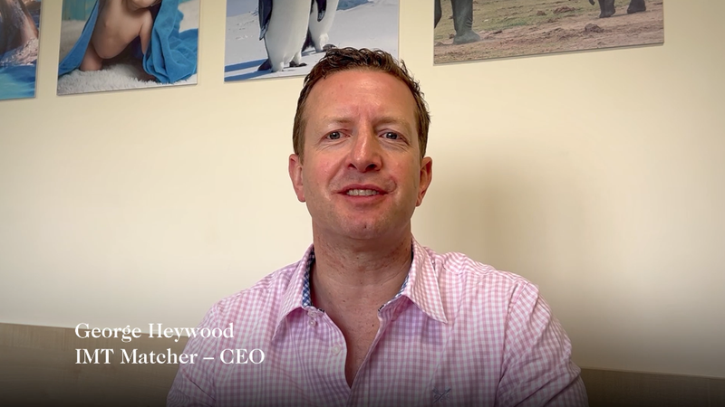 A man in a pink check shirt sitting in front of a beige wall with framed pictures, smiling at the camera. Text overlay: "George Heywood, IMT Matcher – CEO".