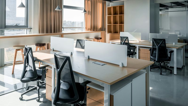 An empty modern office with desks separated by white partitions, black ergonomic chairs, large windows with beige curtains, and wooden storage units.
