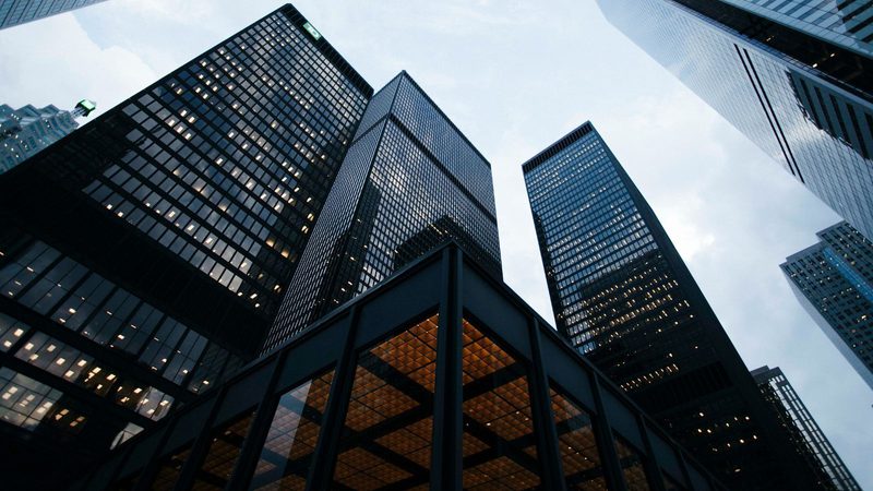 A cluster of tall, modern glass skyscrapers seen from below, reflecting the cloudy sky, in an urban business district.