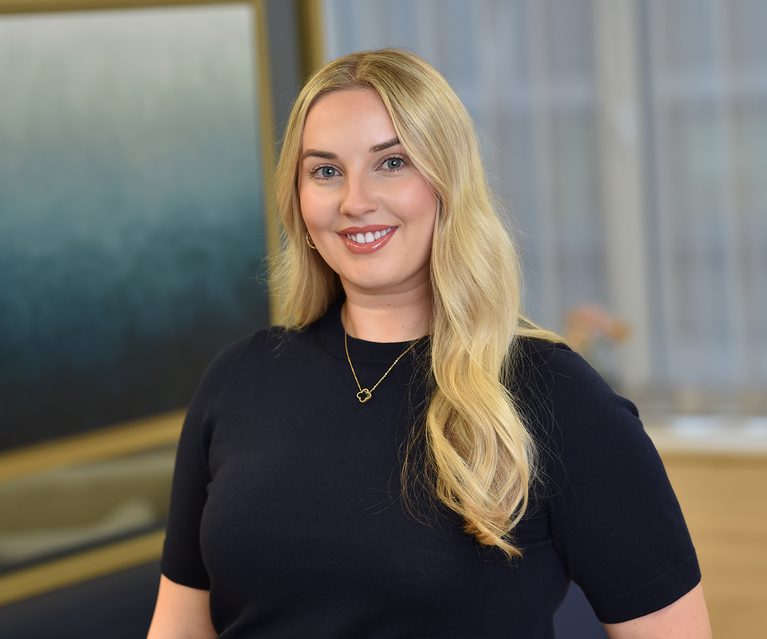 A smiling blonde woman with wavy hair wearing a black top and a gold necklace, standing indoors with a blurred background of windows and modern decor.