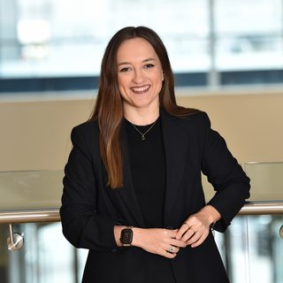A woman with long brown hair, wearing a black blazer and black top, smiling while standing indoors against a glass railing with a modern office background.