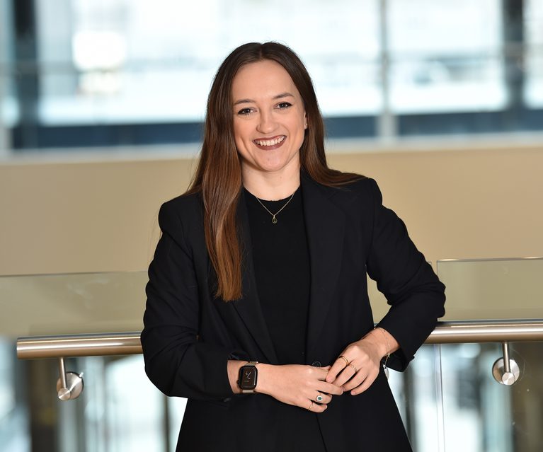 A woman with long brown hair, wearing a black blazer and black top, smiling while standing indoors against a glass railing with a modern office background.