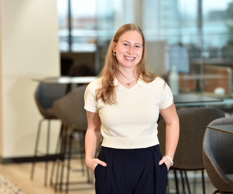 A smiling woman with long blonde hair, wearing a cream short-sleeved top and dark trousers, standing with hands in pockets in a modern office environment.