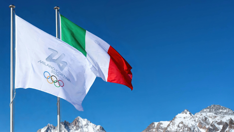 Three flags flying against a blue sky with snow-capped mountains in the background: the Italian flag, the Olympic flag for Milano-Cortina 2026, with the mountain peaks behind.