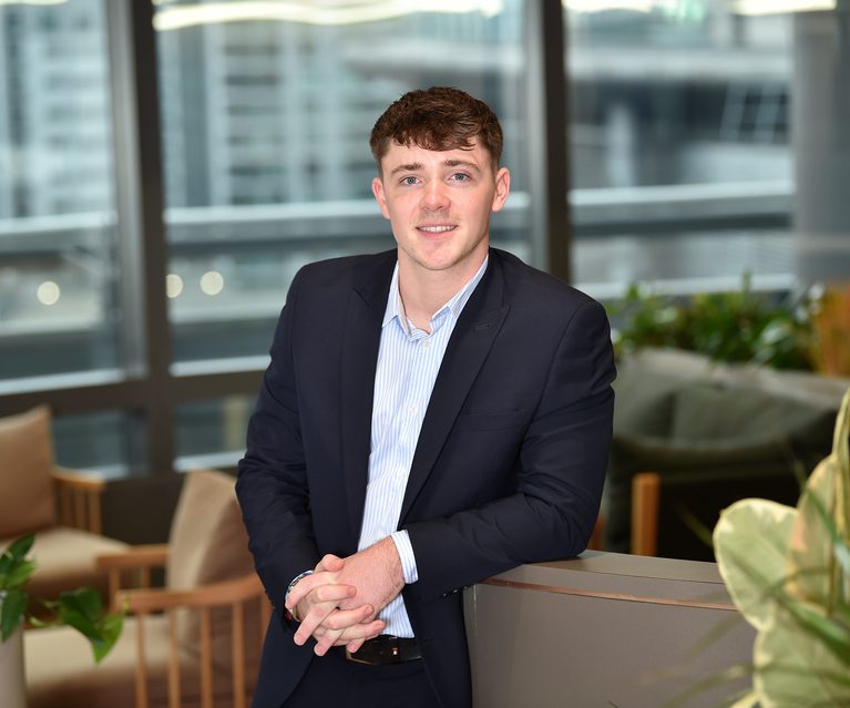 A young man in a dark suit and light blue shirt stands in a modern office, leaning on a desk with large windows and cityscape in the background.