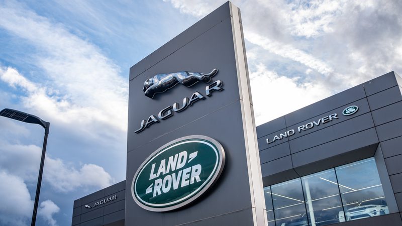 A tall grey sign with Jaguar and Land Rover logos outside a dealership building under a partly cloudy sky.