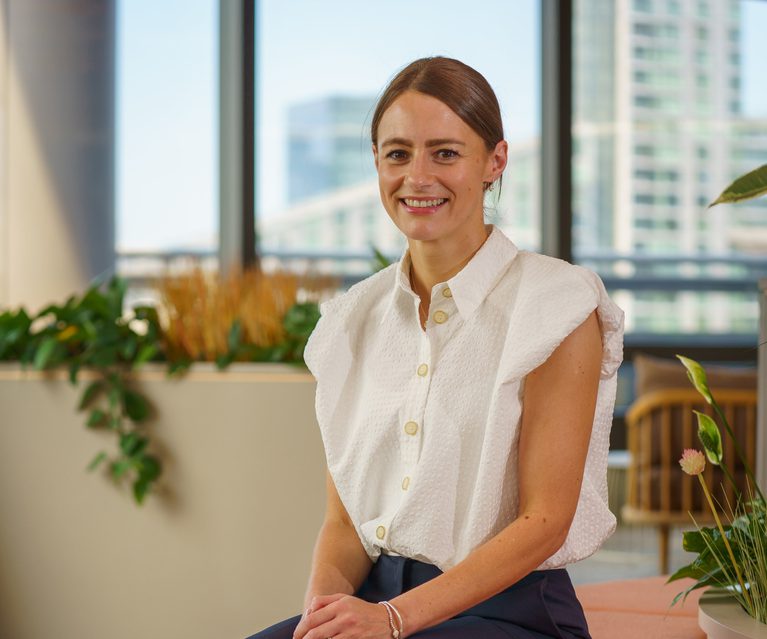 Person seated on a peach lounge sofa in a bright office lobby with plants, wearing a white textured blouse, navy trousers and black heels, smiling at the camera.
