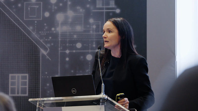 A woman with dark hair and wearing a black blazer is giving a presentation behind a clear podium, with a laptop and a microphone, in front of a technological background.