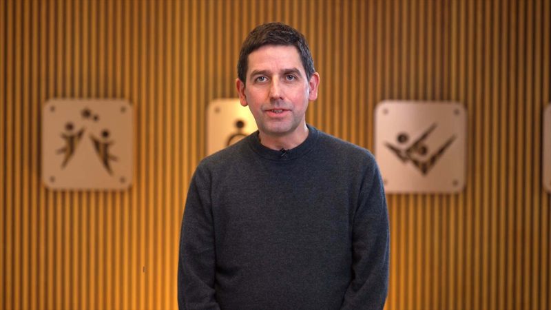 A man with short dark hair and a dark sweater stands in front of a wooden panel wall with abstract art pieces featuring human figures in dynamic poses.