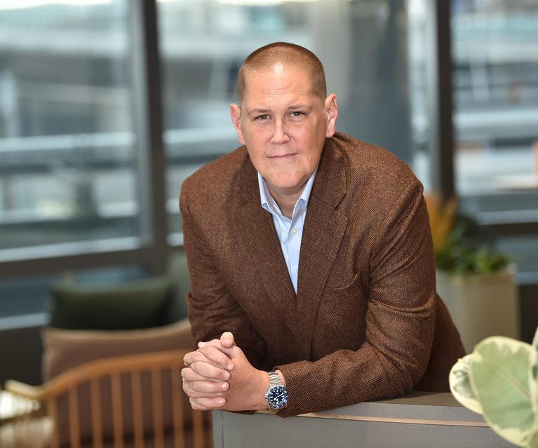 A man with a shaved head wearing a brown blazer and light blue shirt, leaning on a table with hands clasped, in an office environment with large windows behind him.